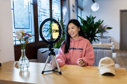 millennial girl brunette hair wearing trendy fashionable casual clothing and making photo or video content for social media with smartphone on tripod an ring lamp